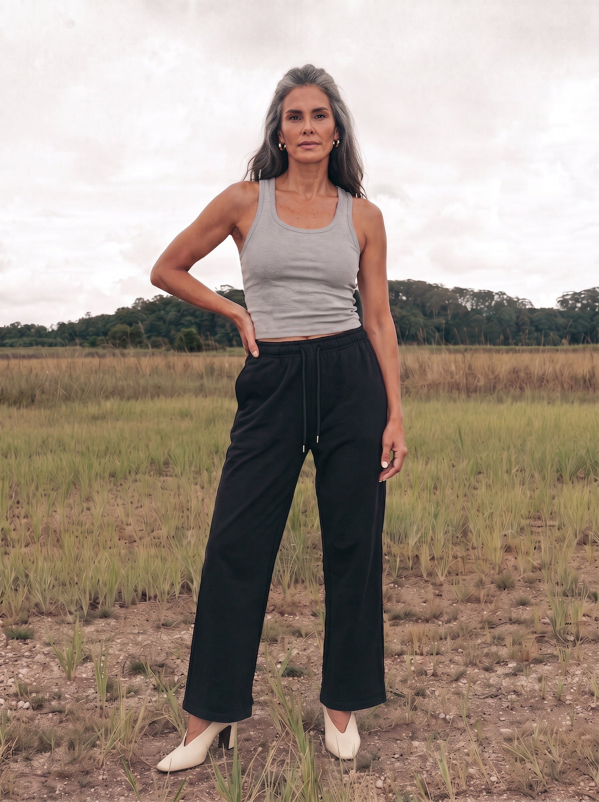 A woman stands outdoors in a grassy field wearing black straight-leg pants, a light gray tank top, and white pointed-toe heels.