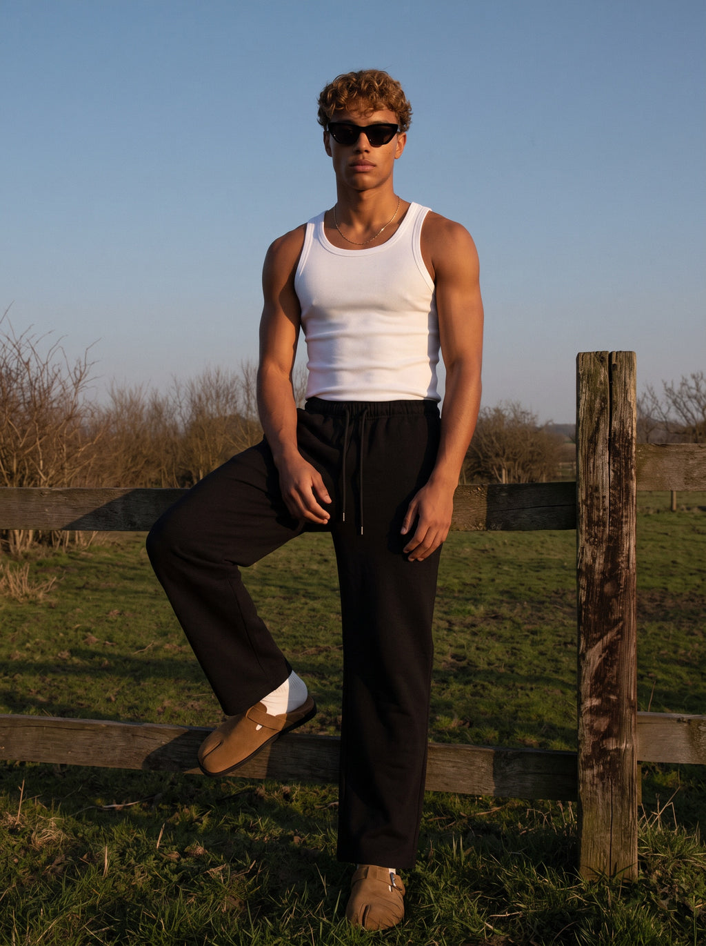 A man wearing a white tank top and black drawstring sweatpants stands outdoors on a grassy field beside a wooden fence, wearing sunglasses and tan loafers.