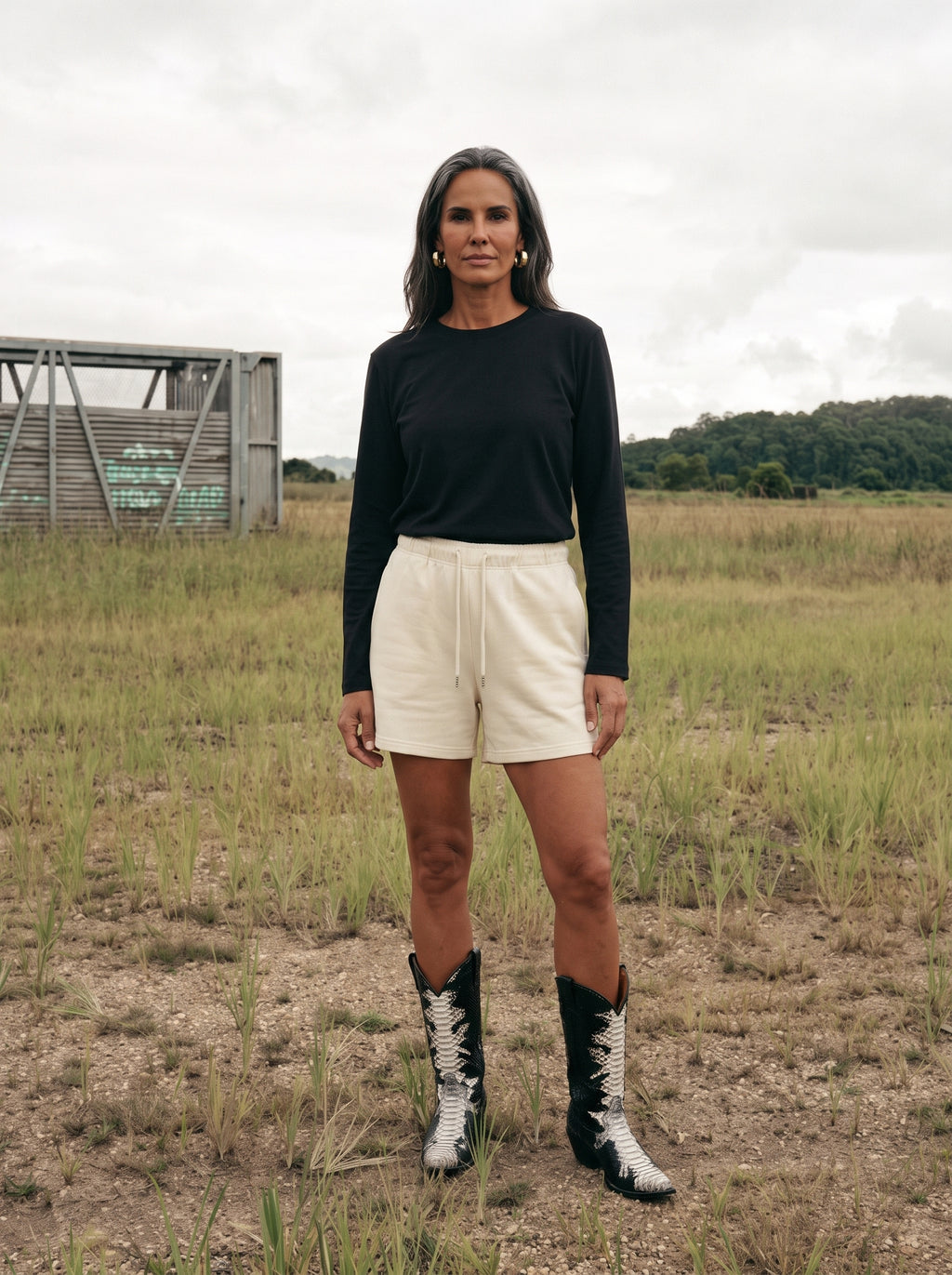 A woman stands outdoors in a grassy field wearing a black long-sleeve top, off-white drawstring shorts, and black-and-white patterned cowboy boots.