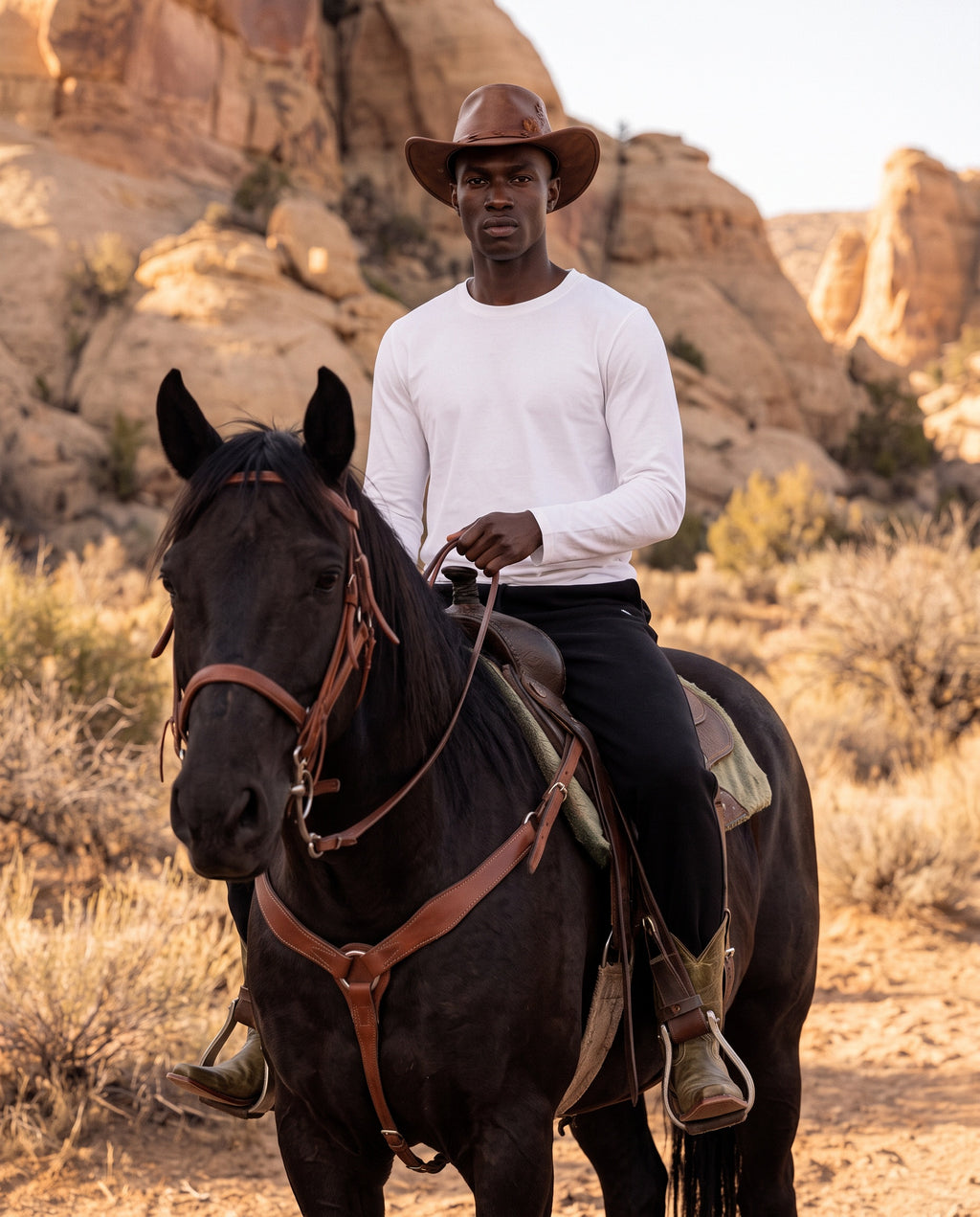 A man wearing a white long-sleeve shirt, black pants, and a brown cowboy hat rides a dark horse through a desert landscape with rock formations in the background.